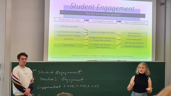Ein Mann und eine Frau stehen vor einer Tafel. Über der Tafel wird eine Präsentation angezeigt. Die Überschrift der Präsentation lautet Student Engagement.