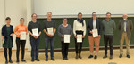 Neun Personen stehen nebeneinander auf einer Bühne. Sechs halten eine Urkunde in der Hand.