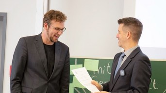 Zwei Personen lächeln in die Kamera. Die rechte Person hält eine Urkunde zur Übergabe bereit.