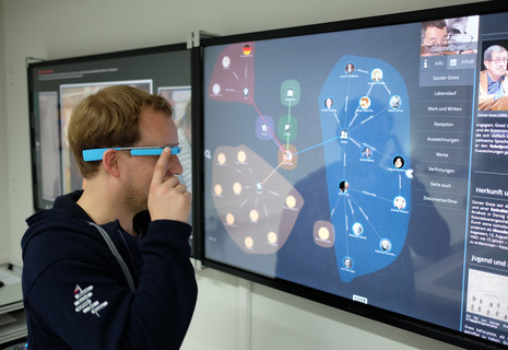 Eine Person steht vor einem Fernseher und bedient mit seinem Finger eine Google Glass Brille